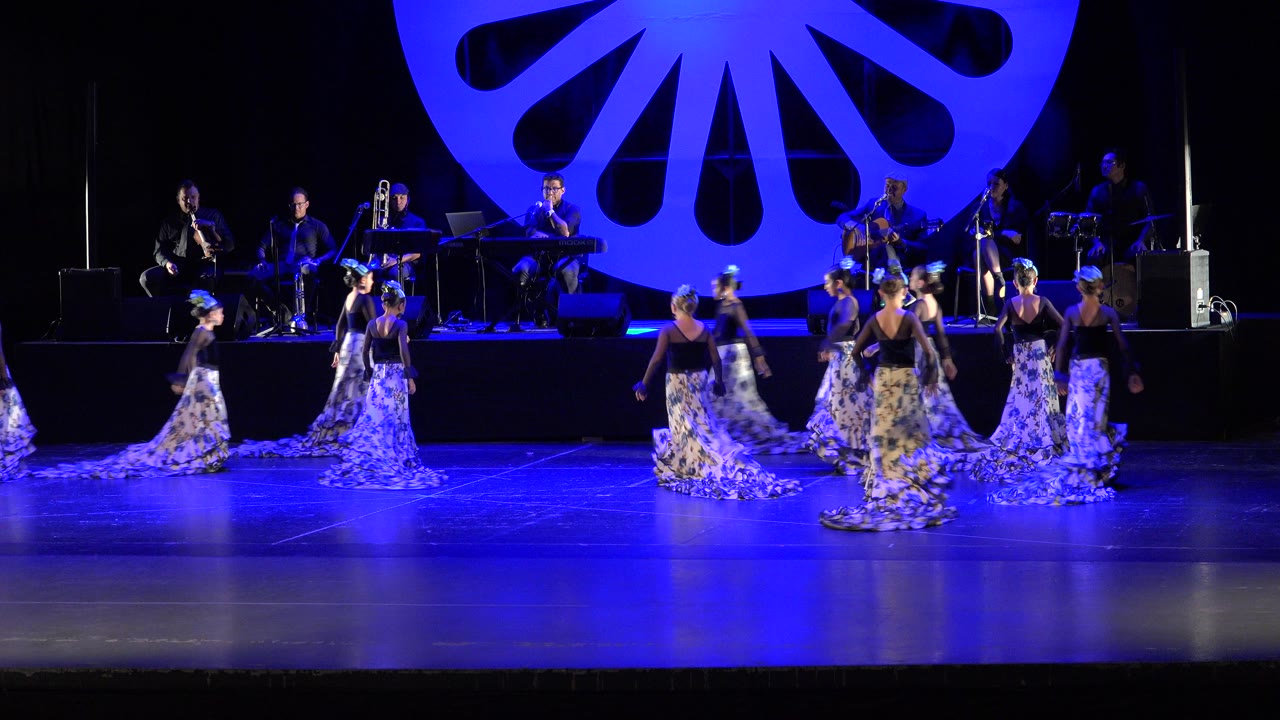 Women in silver-blue skirts dance on a blue-lit stage with musicians behind them.