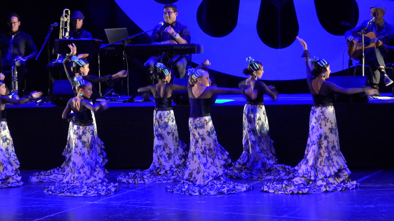 Several young dancers in matching floral skirts hold extended arm positions on the stage while live musicians remain seated in the background in Cabo San Lucas, Mexico.