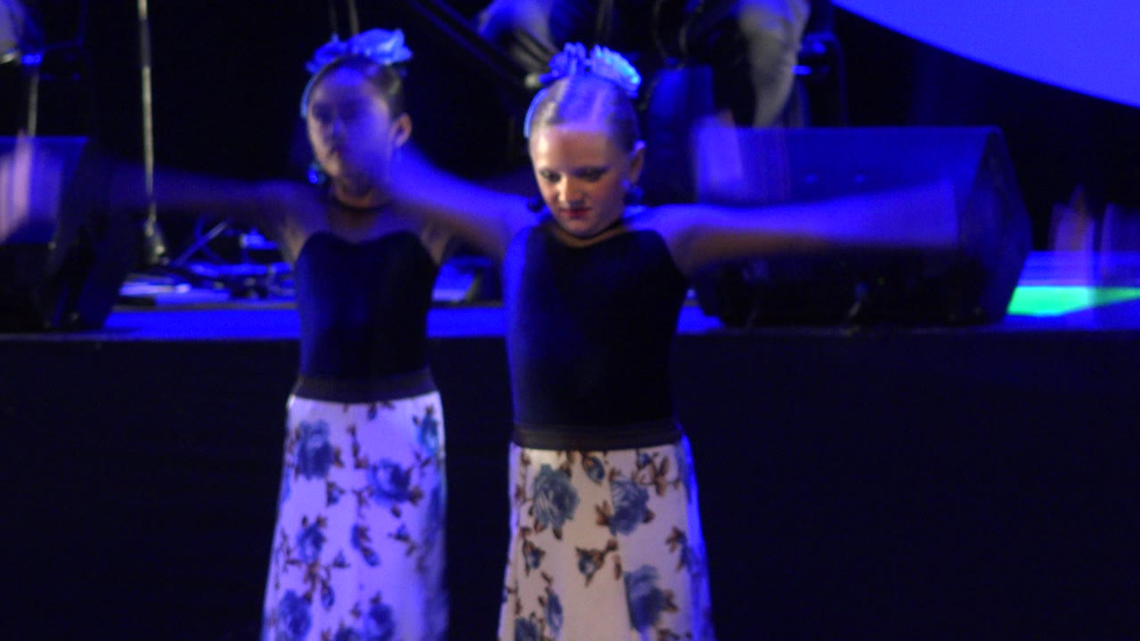 Two girls continue the dance routine on a dark stage in Cabo San Lucas, Mexico, with raised arms, floral pants, and blue lighting across the floor equipment behind them.