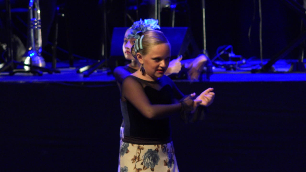 A young girl in a black dance top and floral skirt performs hand gestures on a blue-lit stage in Cabo San Lucas, Mexico, with another child partly visible behind her.
