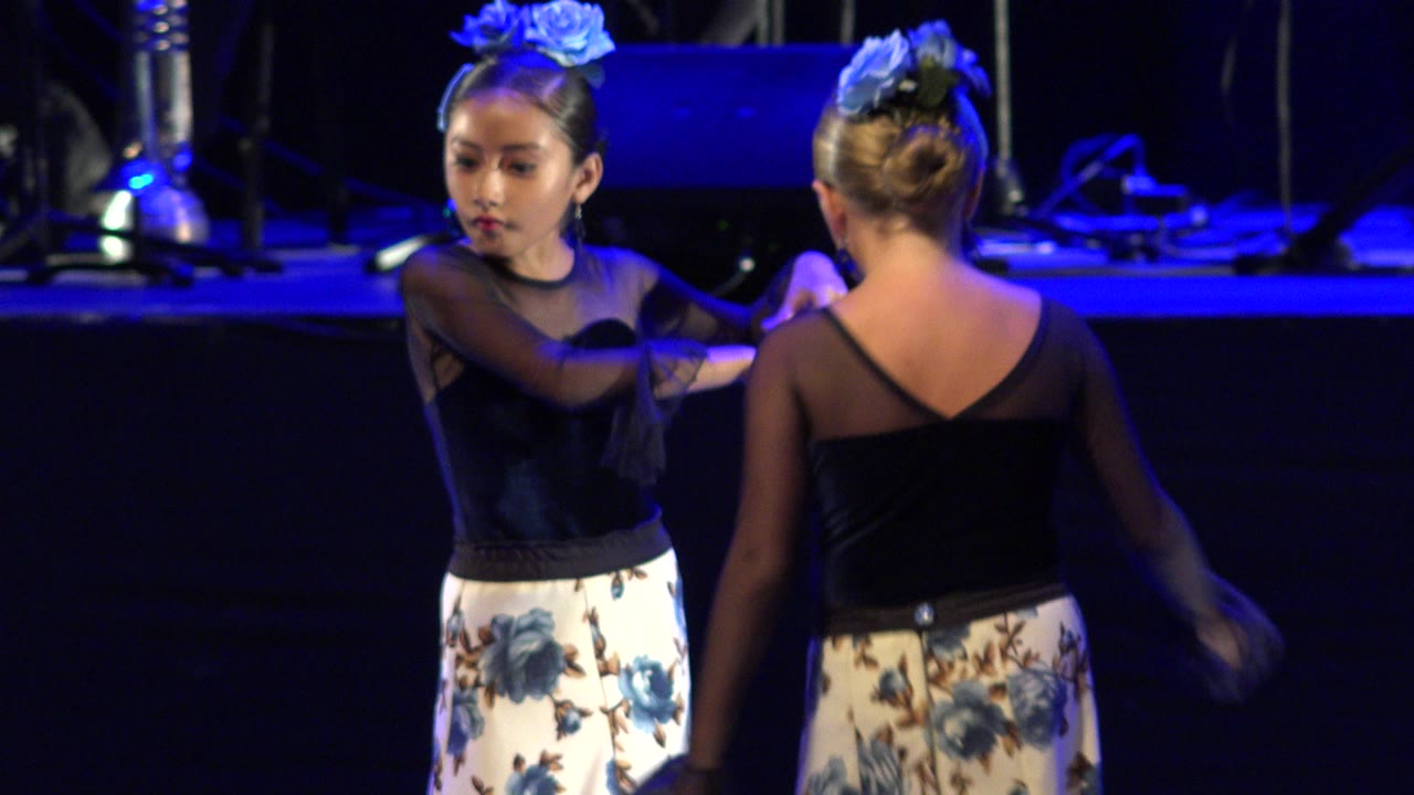 The dancers hold a close-facing position on the Cabo San Lucas stage, with one girl's face visible and the other turned away from the camera.