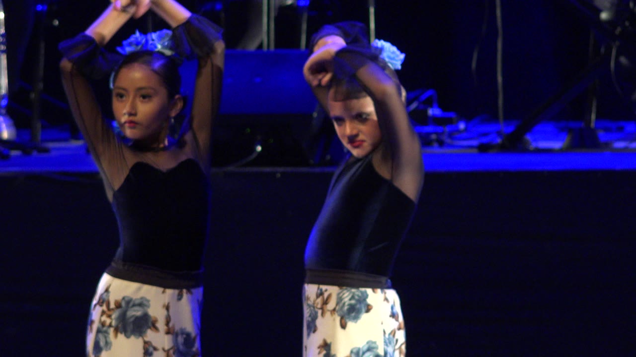 Two young girls in black tops and floral skirts hold raised arm positions on a blue-lit stage during a Cabo San Lucas dance performance.