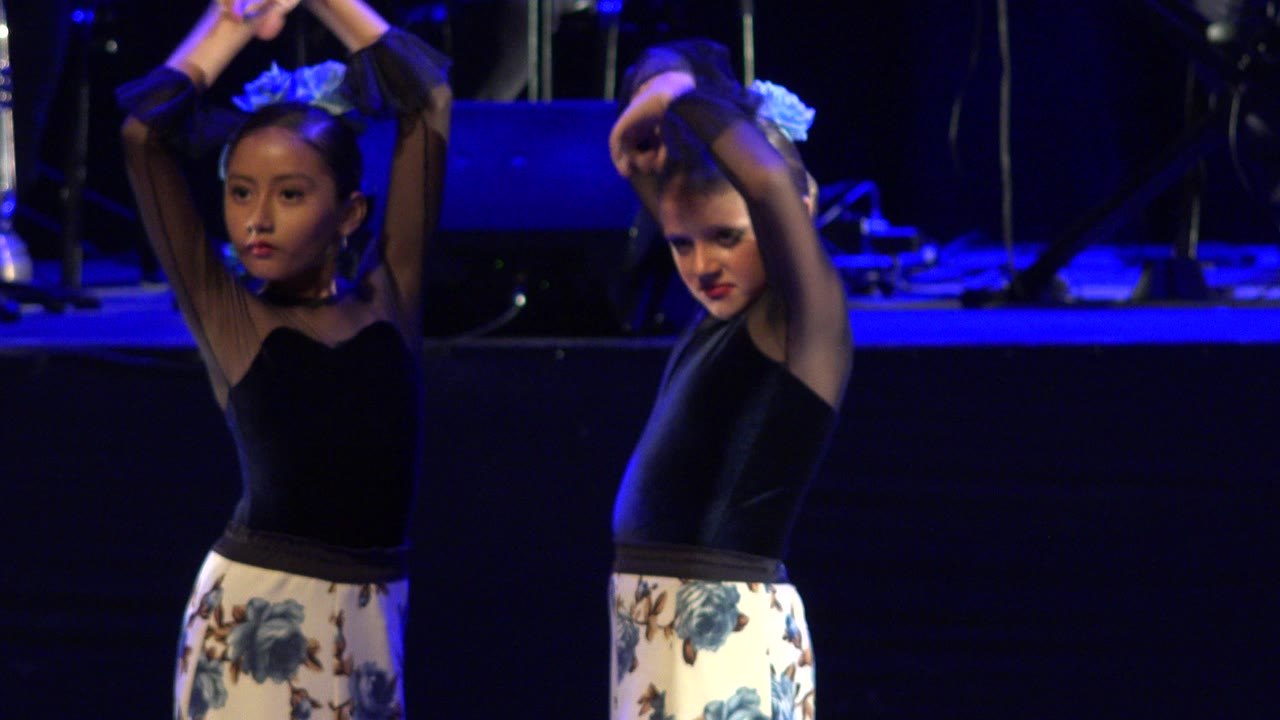 The two child dancers remain side by side with arms lifted, black tops and pale floral skirts visible against the dark Cabo San Lucas stage background.