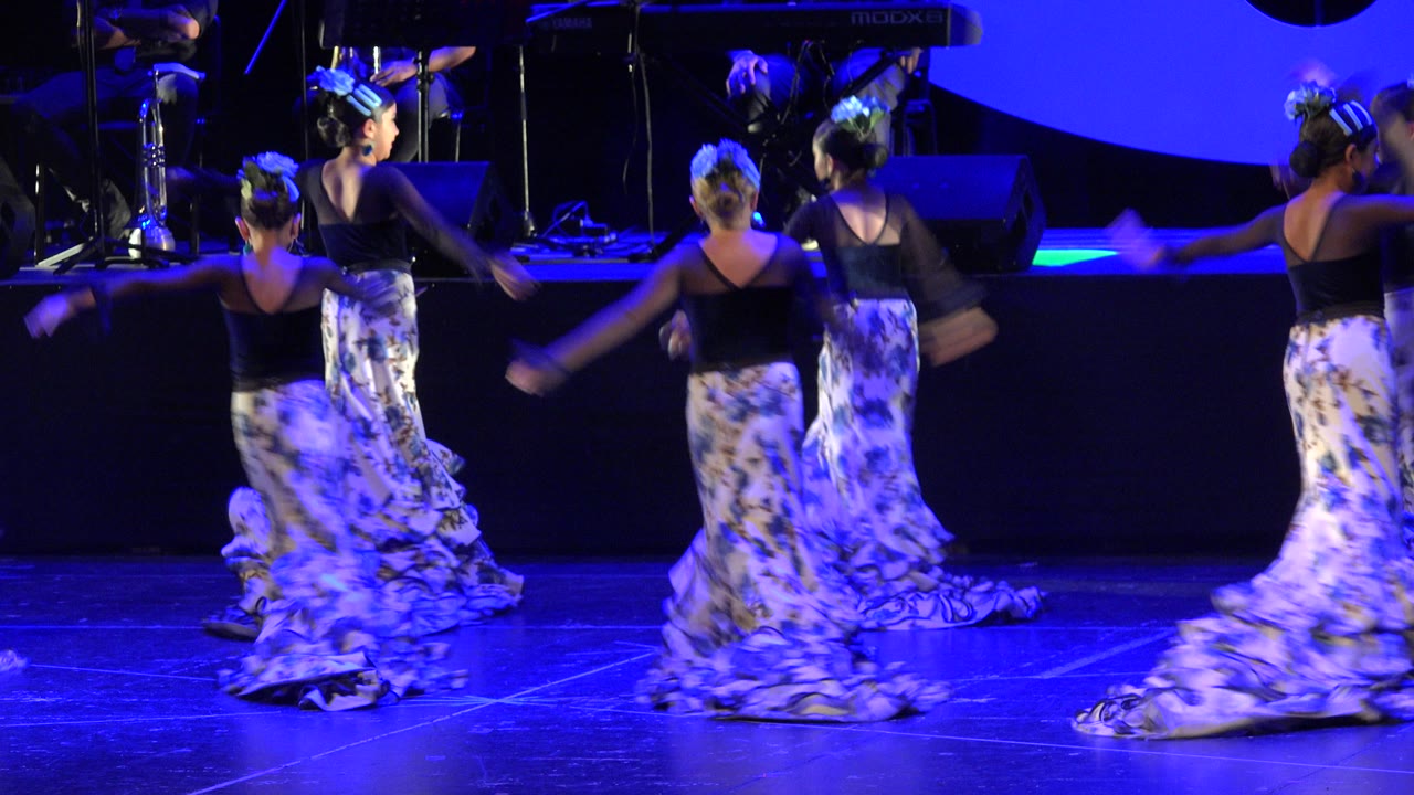 Several young dancers turn across the stage in Cabo San Lucas, Mexico, with floral skirts blurring from movement and black stage equipment behind them.