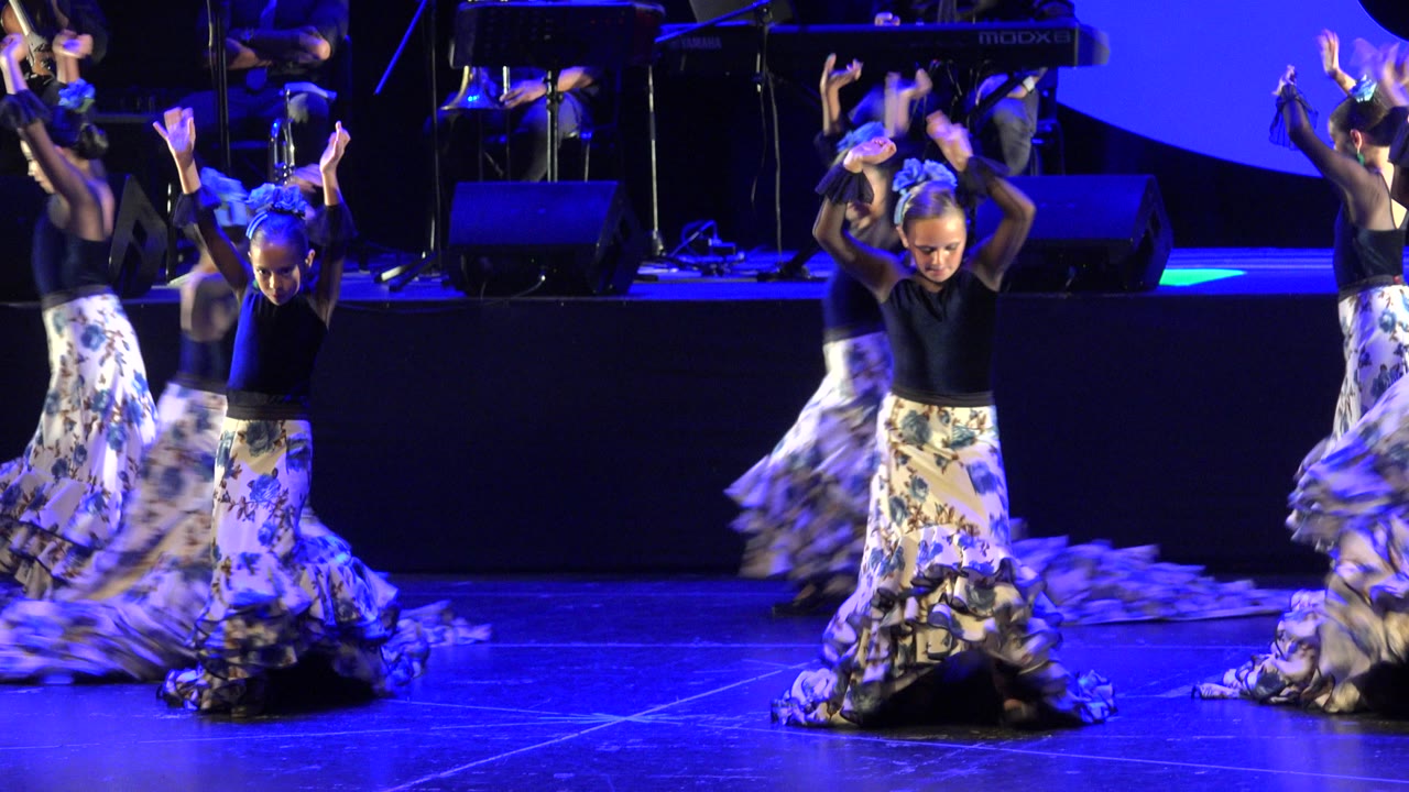 Children perform a choreographed dance on a blue-lit stage in Cabo San Lucas, Mexico, with musicians and equipment visible behind them.