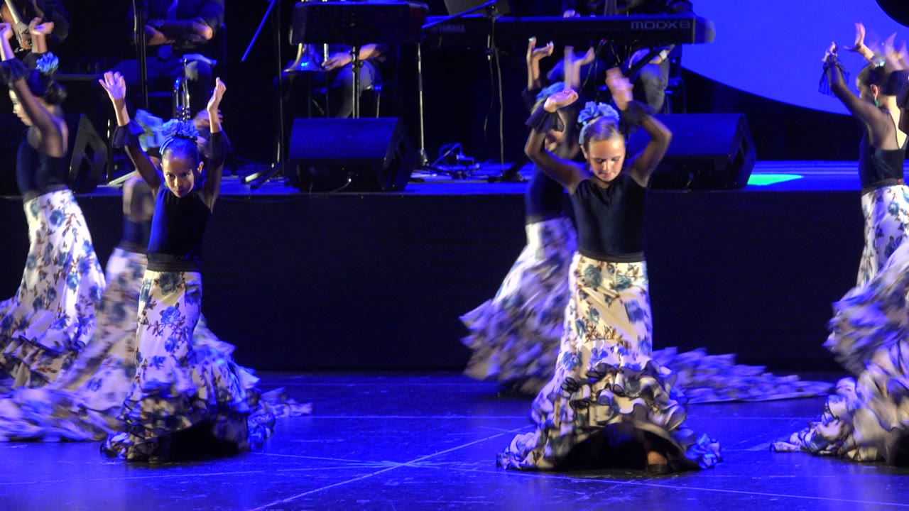 A child dancer stands near center with arms raised while other children blur through the routine on the Cabo San Lucas stage.