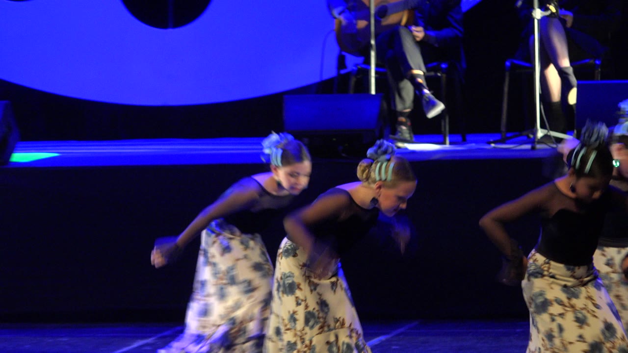 Three girls dancing on a blue-lit stage in floral skirts.