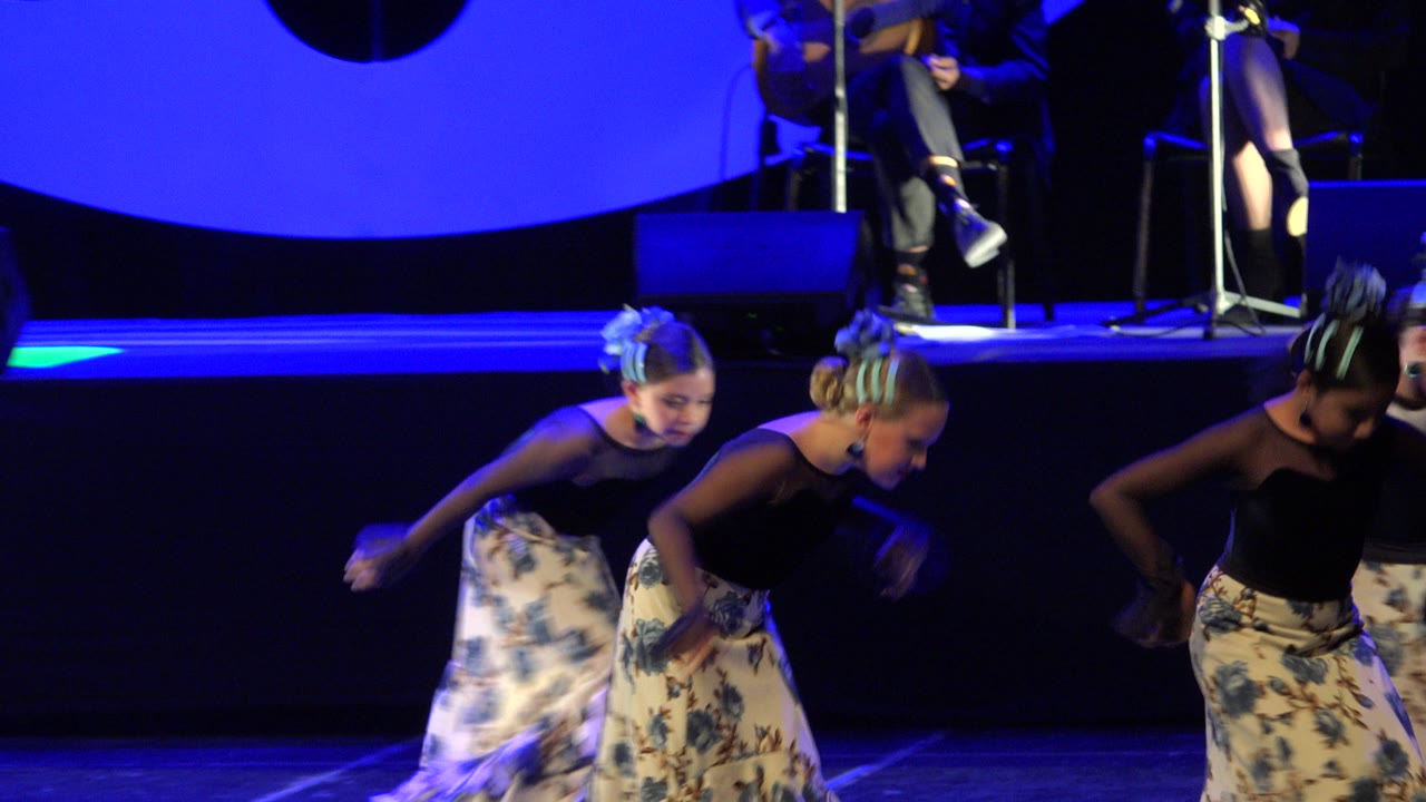 The dancers lean forward in a coordinated step on the blue-lit performance stage in Cabo San Lucas, Mexico, with instruments and seated performers in the background.