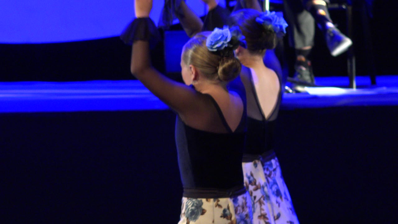 Two young girls continue the stage routine in Cabo San Lucas, Mexico, with one dancer's arm raised and another turned slightly away under blue lighting.