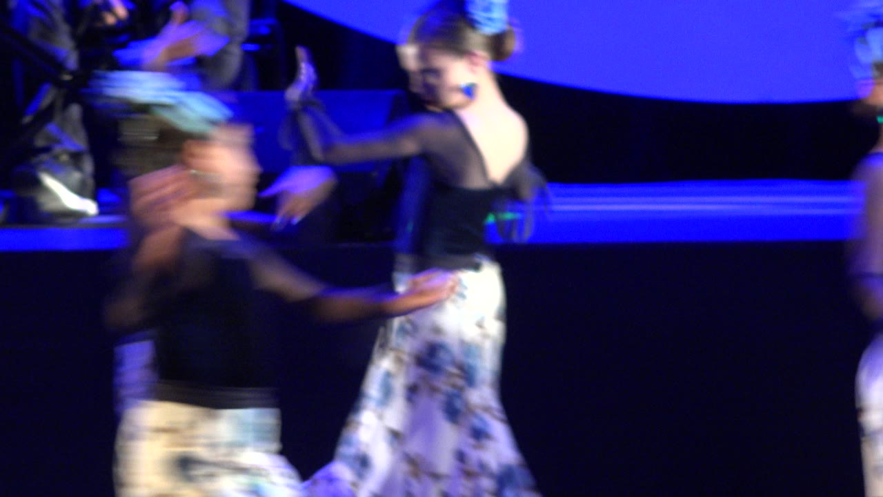 Several young dancers pass in front of a raised band area in Cabo San Lucas, Mexico, with blue light washing the stage and a keyboard player in the background.