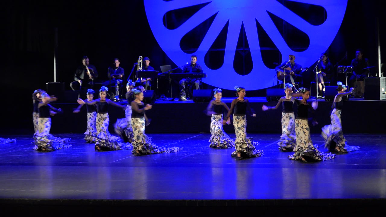 Wide stage with dancers in patterned skirts and musicians behind them under blue lighting.