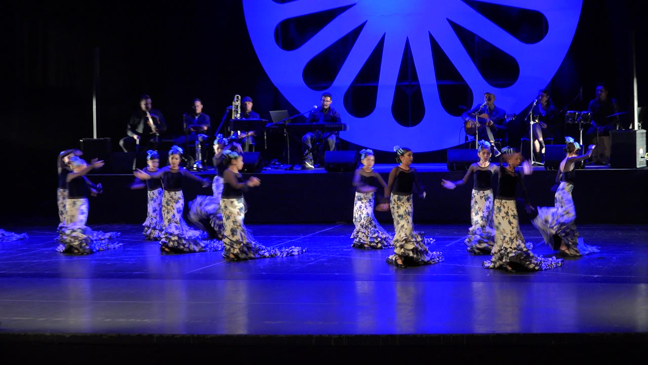 Cabo San Lucas dancers move through a wide formation at center stage, with several performers facing different directions and musicians visible behind them in blue light.