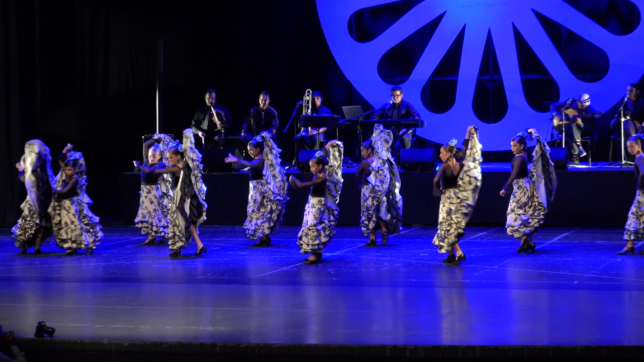 Girls in patterned skirts dance on a blue-lit stage with musicians behind them.