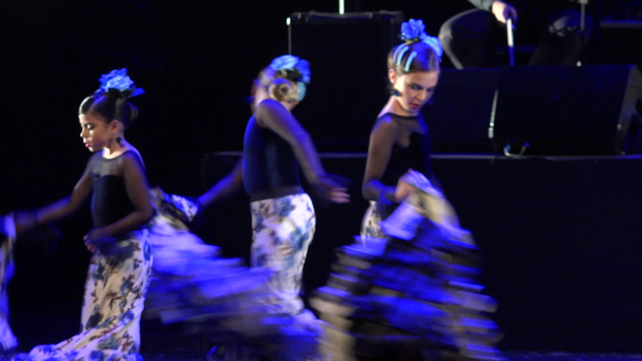 Three young girls perform on a dark stage in Cabo San Lucas, Mexico, with one dancer at left and two girls crossing at center under blue light.