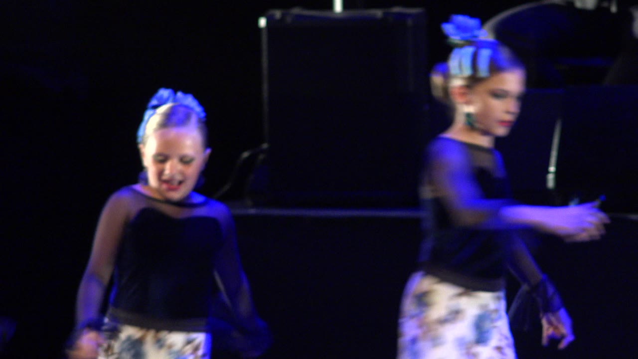 Child dancer under blue stage light in a black top and white floral skirt.