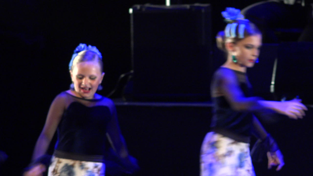 Two child dancers in Cabo San Lucas, Mexico appear on a dark stage, with the foreground dancer moving quickly under blue light.
