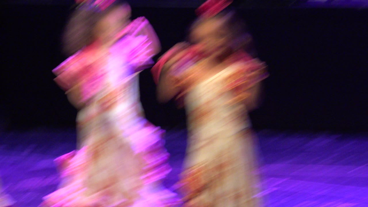 Two young dancers in matching light costumes are blurred by motion as they move across the dark stage in the Cabo San Lucas performance.