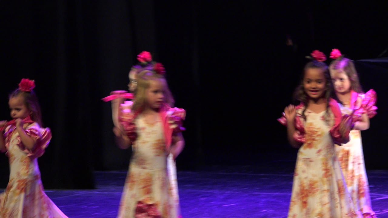 Young girls in pink and yellow dresses dancing on a dark stage.
