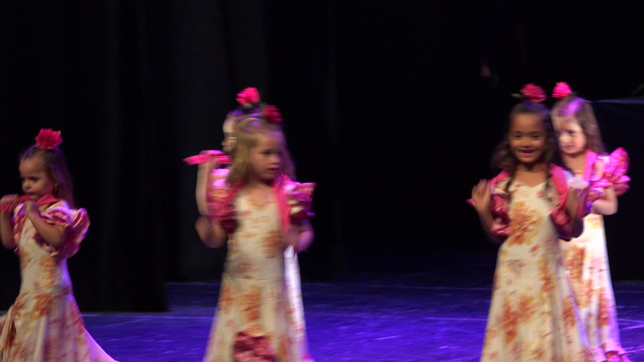 Several young girls continue the stage dance in Cabo San Lucas, Mexico, with one child blurred in the foreground and others lined across the dark curtained background.