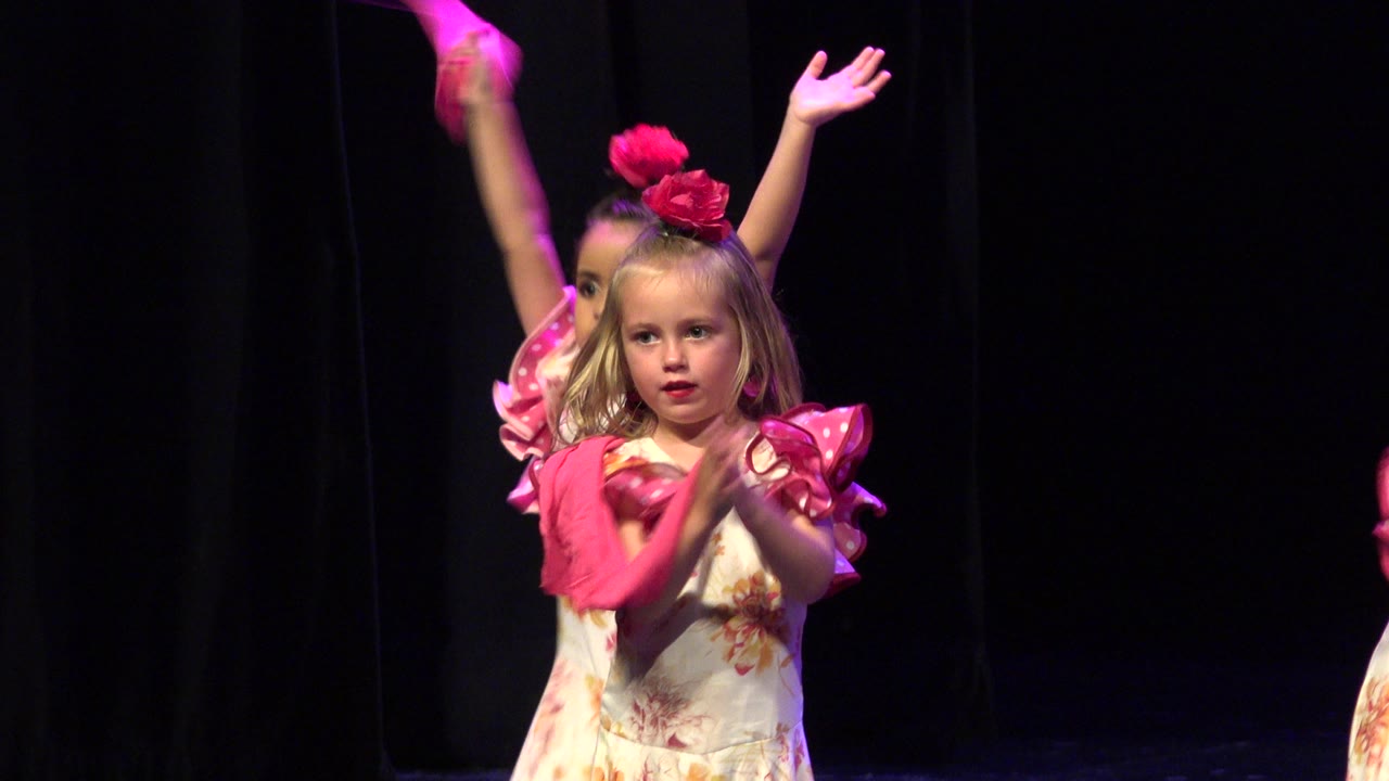 A preschool-aged girl in Cabo San Lucas wears a floral dress with pink ruffled sleeves and a pink headpiece while clapping under stage light, with another child partly visible behind her.