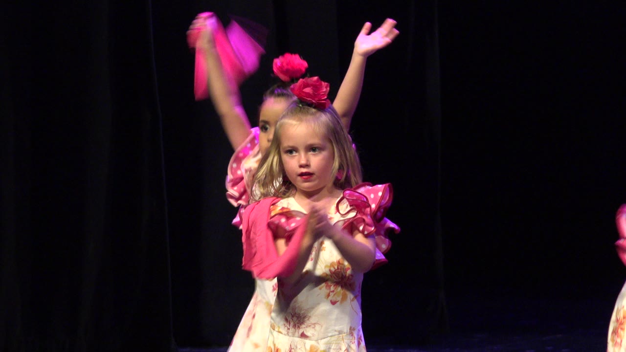 The child performer remains centered in Cabo San Lucas, clapping in a floral costume while a bright pink scarf from another dancer streaks through the dark stage background.