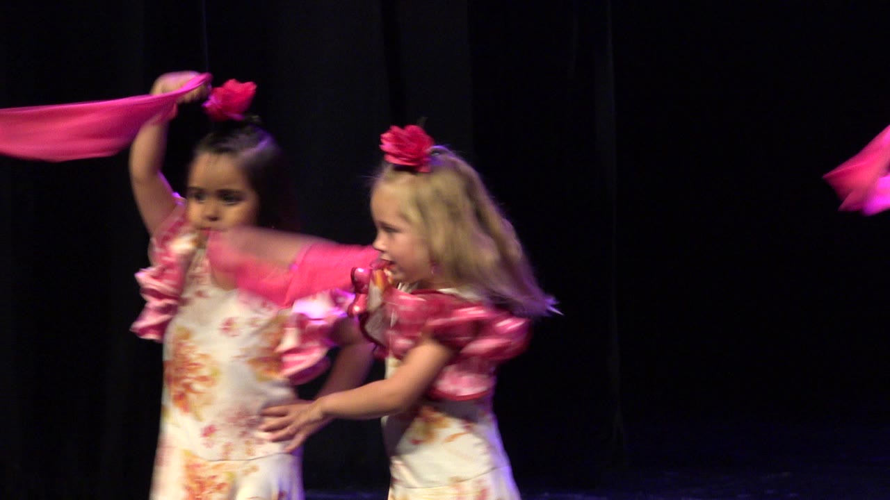 Two young girls in floral dance costumes on a dark stage with a pink scarf