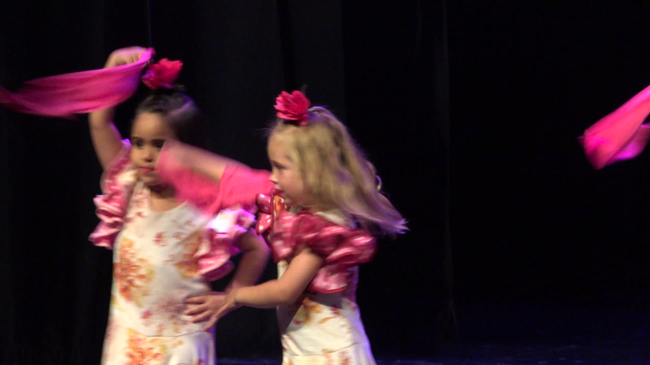 The Cabo San Lucas stage performance continues as a girl raises a bright pink scarf while another child in a matching floral costume stands beside her.