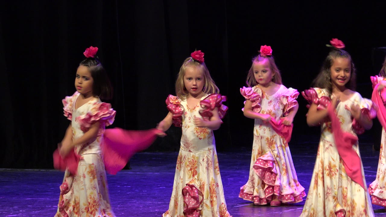 Girls in floral ruffled dresses stand on stage with pink shawls during the routine.