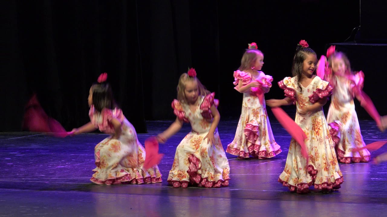 Young girls dancing with pink ribbons on a dark stage.
