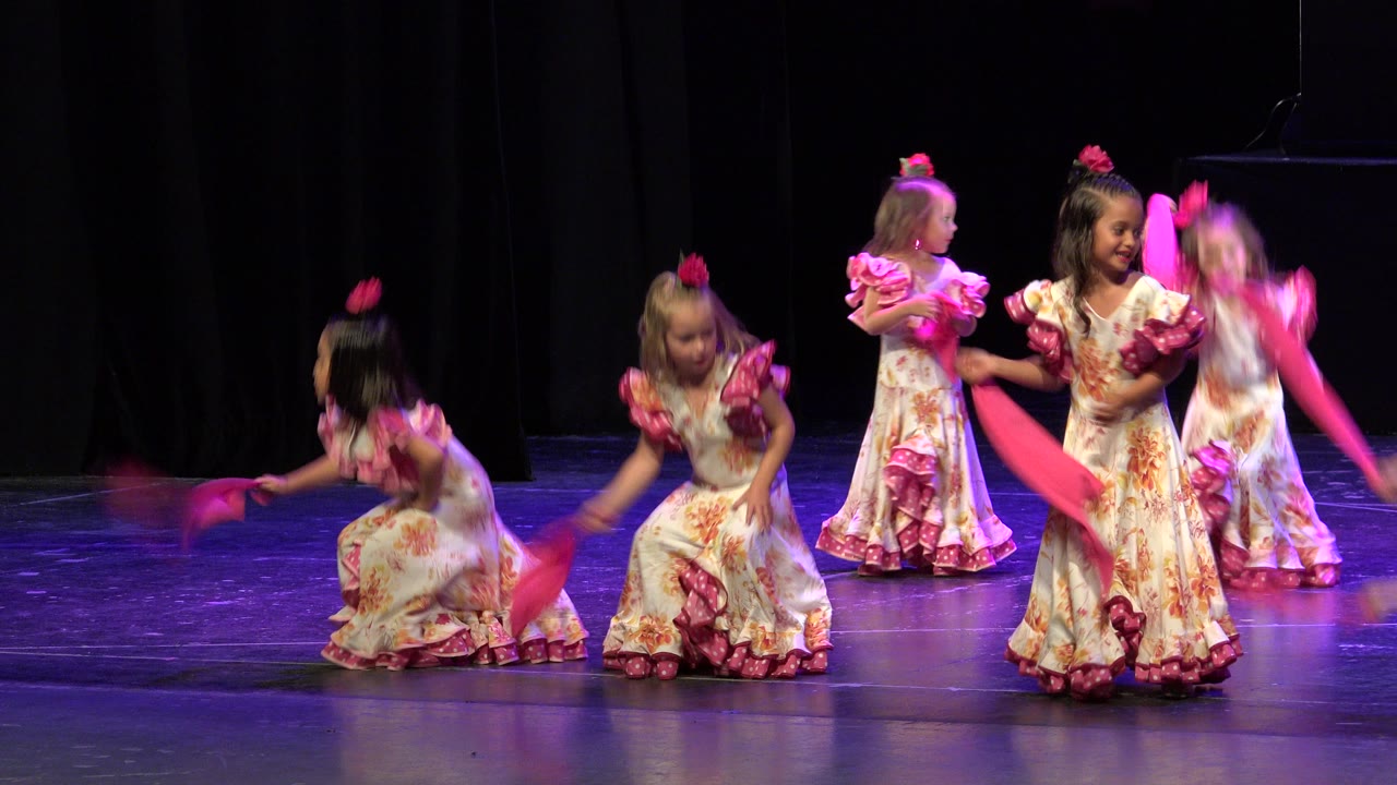 Several children in matching floral dresses continue the flamenco-style stage routine, with one dancer crouched and others standing behind her.