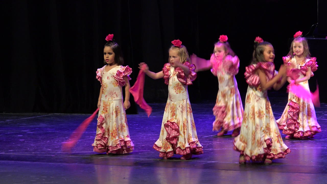 Girls in pink and white dresses dance on a dark stage with scarves.