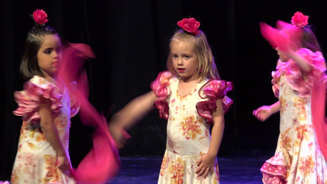 A young girl in a floral dress and pink hair flower waves a bright pink scarf on a dark stage in Cabo San Lucas, Mexico.