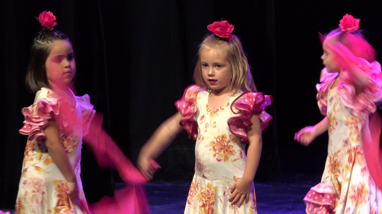 A blonde young girl in a floral dress faces the audience while another child in matching costume turns behind her on the Cabo San Lucas stage.