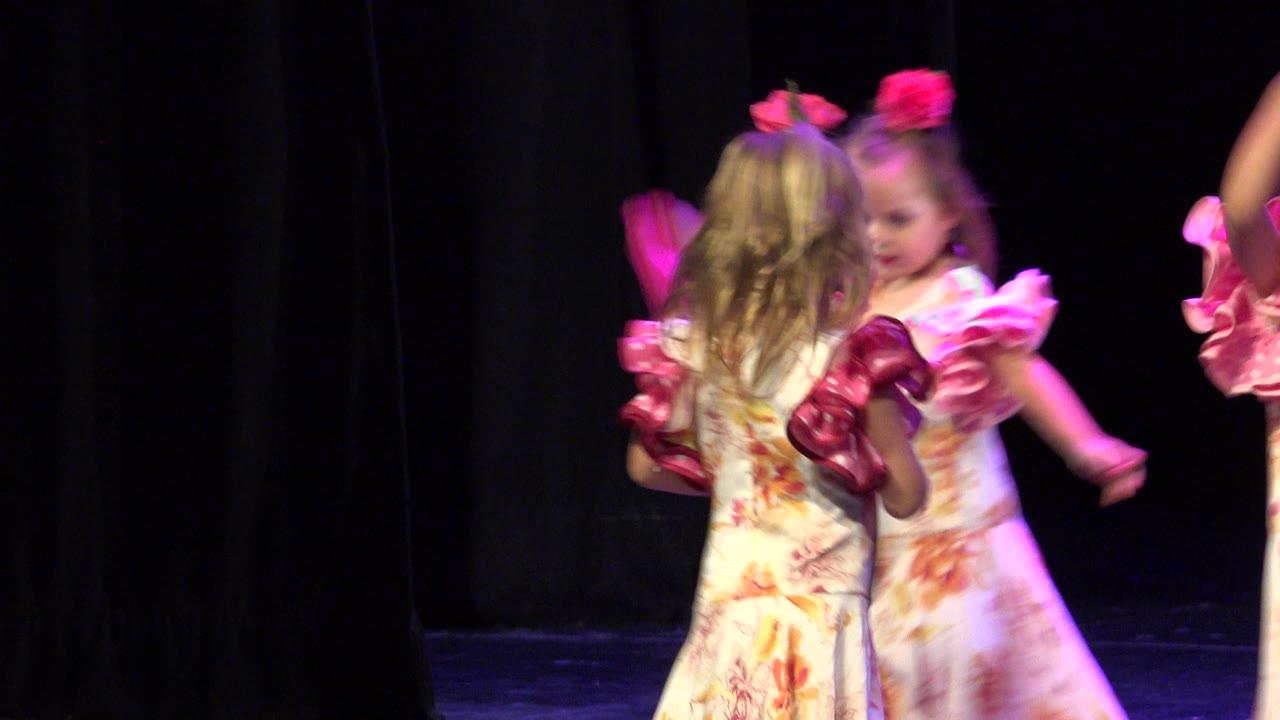 Two young girls in floral dance dresses stand close together on a dark stage in Cabo San Lucas, with pink ruffles and hair flowers lit by magenta stage light.