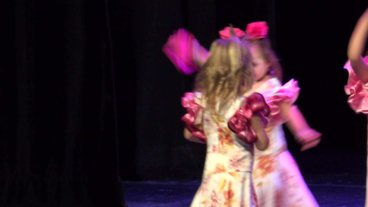 Young girls in matching floral costumes shift on the Cabo San Lucas stage while bright pink fabric moves through the foreground and edge of frame.