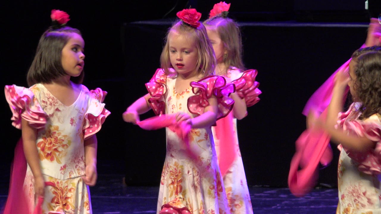 Young girls in floral costumes stand on a dark stage with pink scarves.