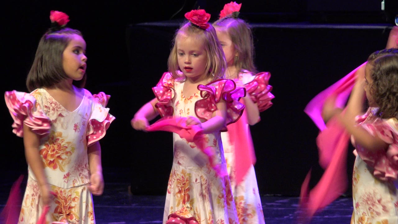 In Cabo San Lucas, Mexico, several young girls in matching floral costumes face inward on stage while pink scarves move at the edge of the frame.