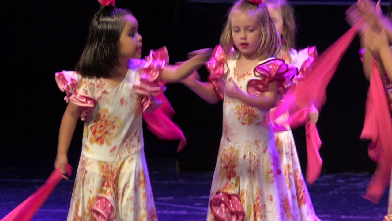 Two little girls in floral dresses hold pink scarves on a dark stage.