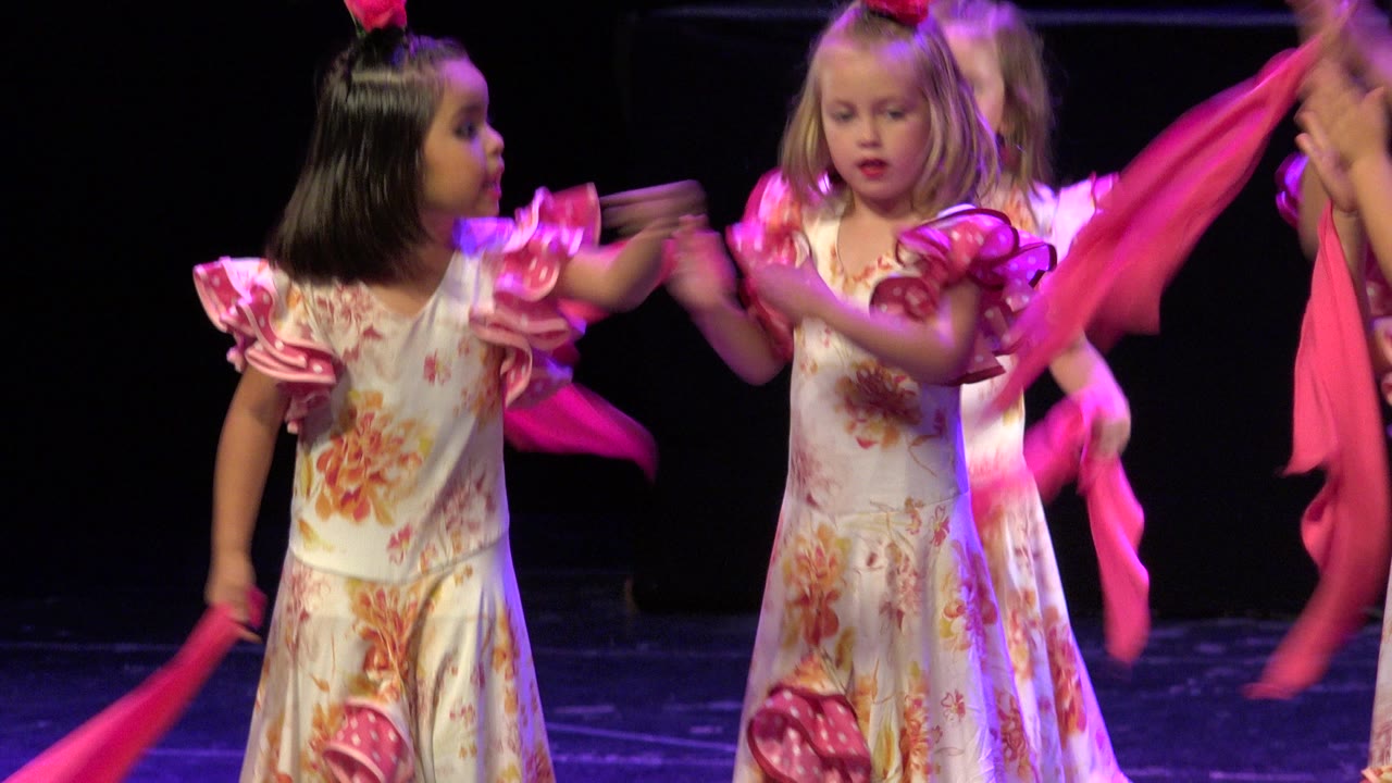 Two young dancers in matching floral costumes face forward and sideways as pink scarves pass through the foreground on the dark Cabo San Lucas stage.