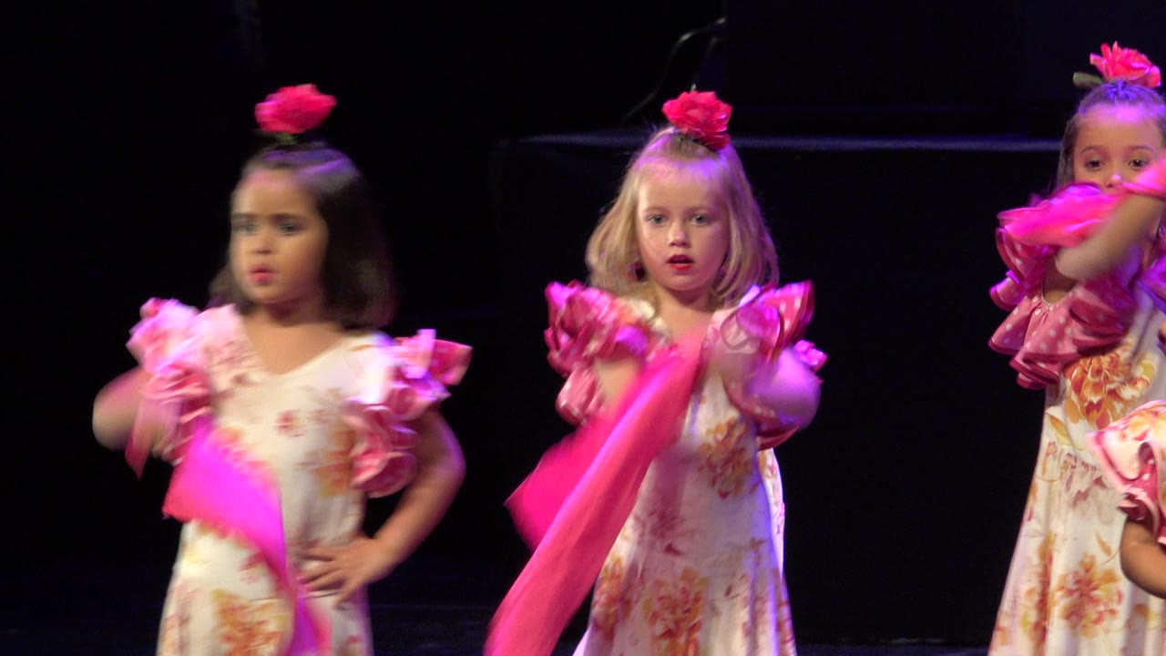 Three young girls in floral cream dresses and pink ruffled sleeves perform with bright pink scarves on a dark stage in a Cabo San Lucas dance clip.