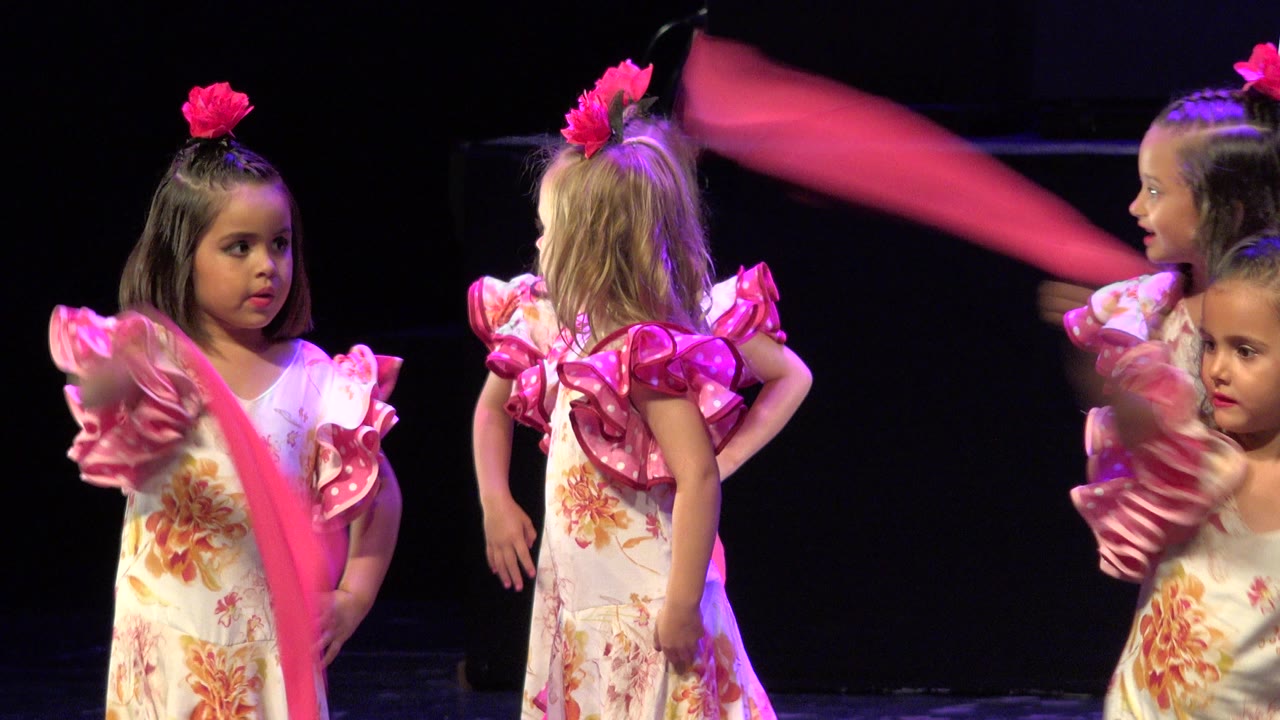 Girls in floral pink dance costumes stand under stage lighting in Cabo San Lucas while a pink scarf blurs across the foreground.