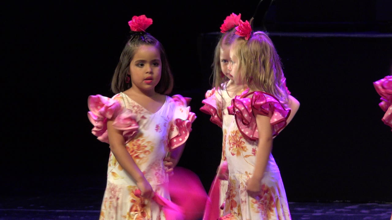 Two young girls in floral dance dresses stand on a dark stage with pink scarves.