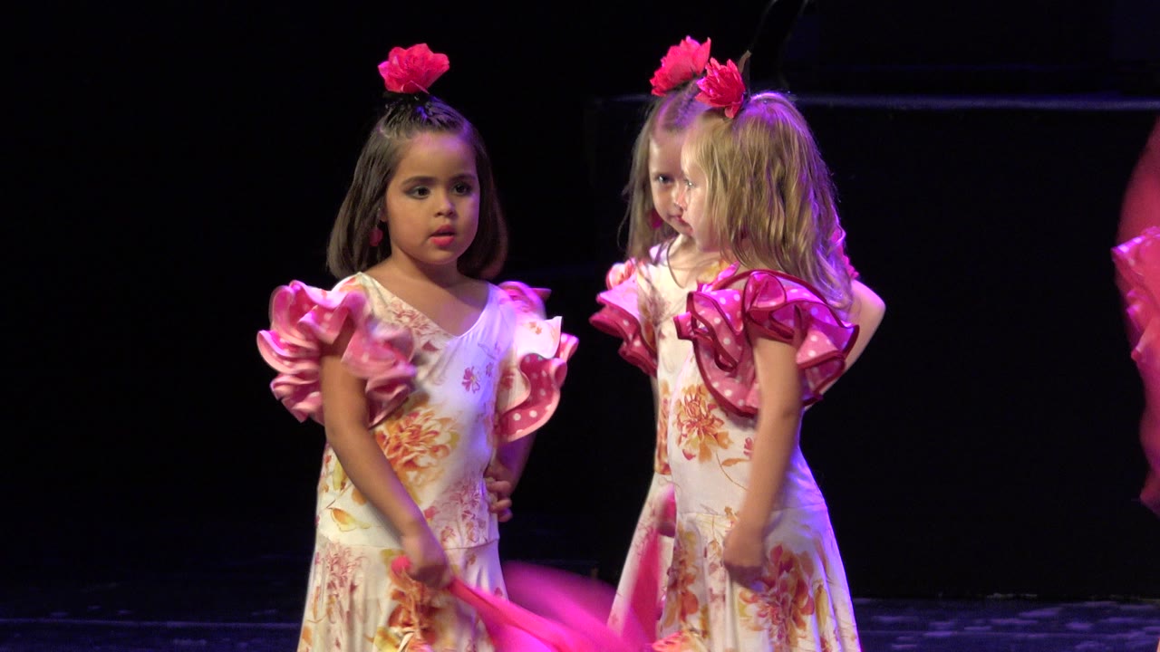 The same two young dancers remain near center on the Cabo San Lucas stage as one pink scarf blurs at the right edge and another crosses low in front.