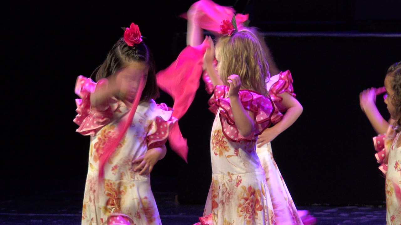 Two young girls in pink floral stage dresses wave scarves on a dark stage.