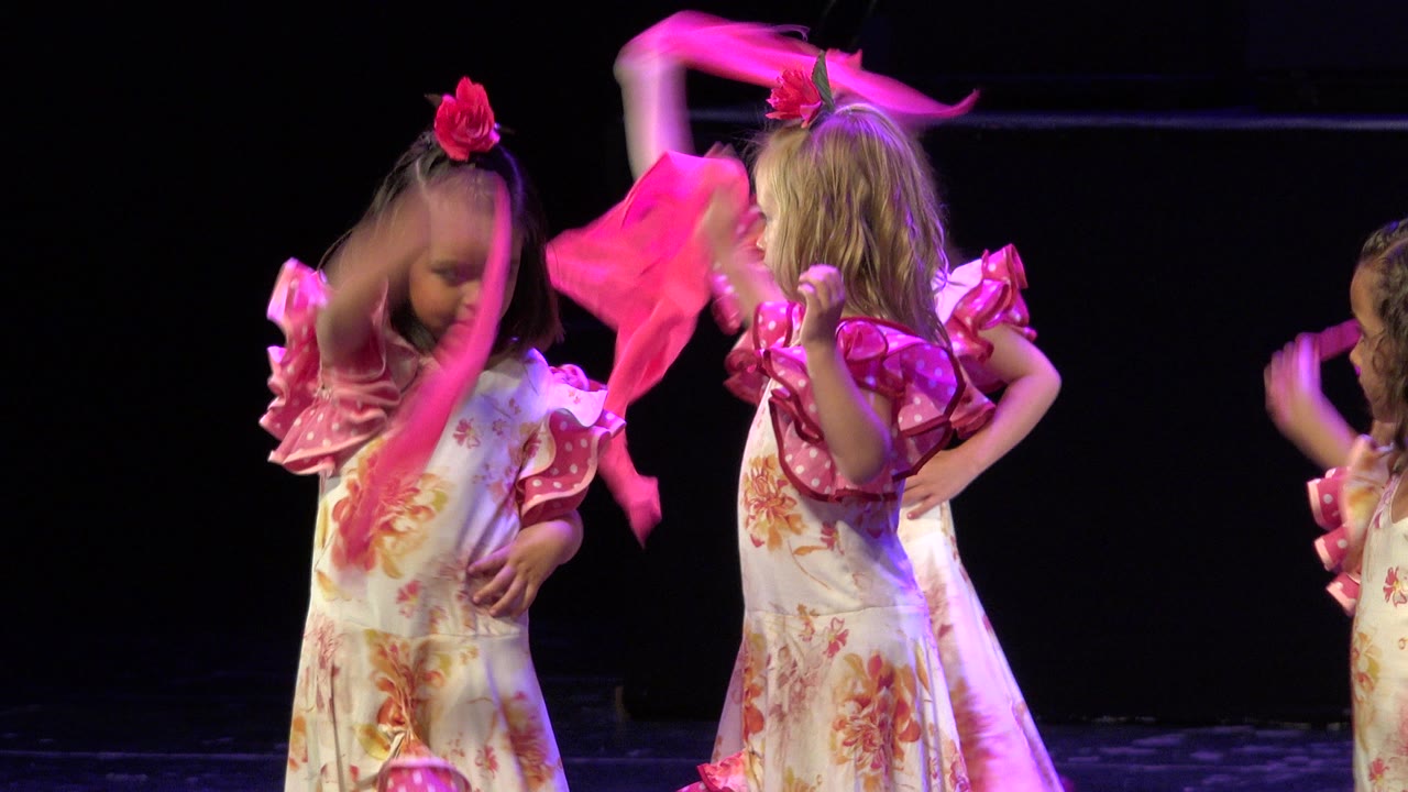 Two young performers in Cabo San Lucas, Mexico stand side by side in pink floral costumes, with one girl's scarf raised overhead and another child partly visible at the right edge.