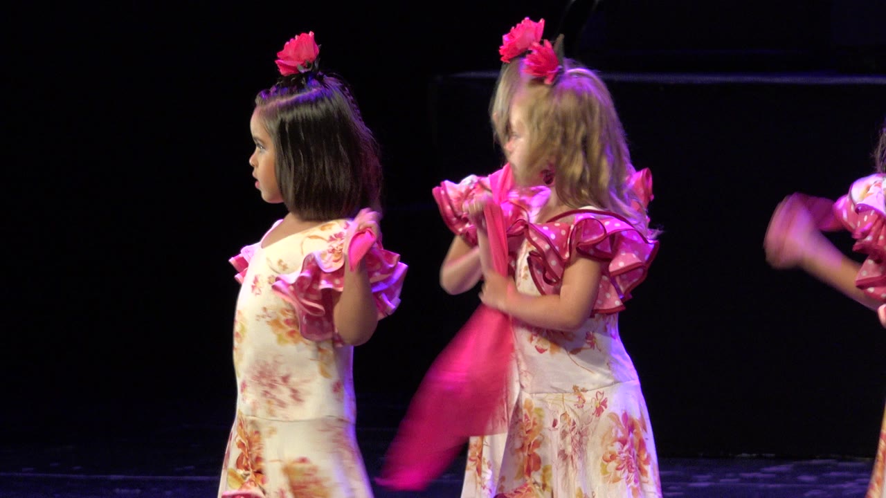 Two young girls in floral dance dresses stand on a dark stage with pink handkerchiefs.