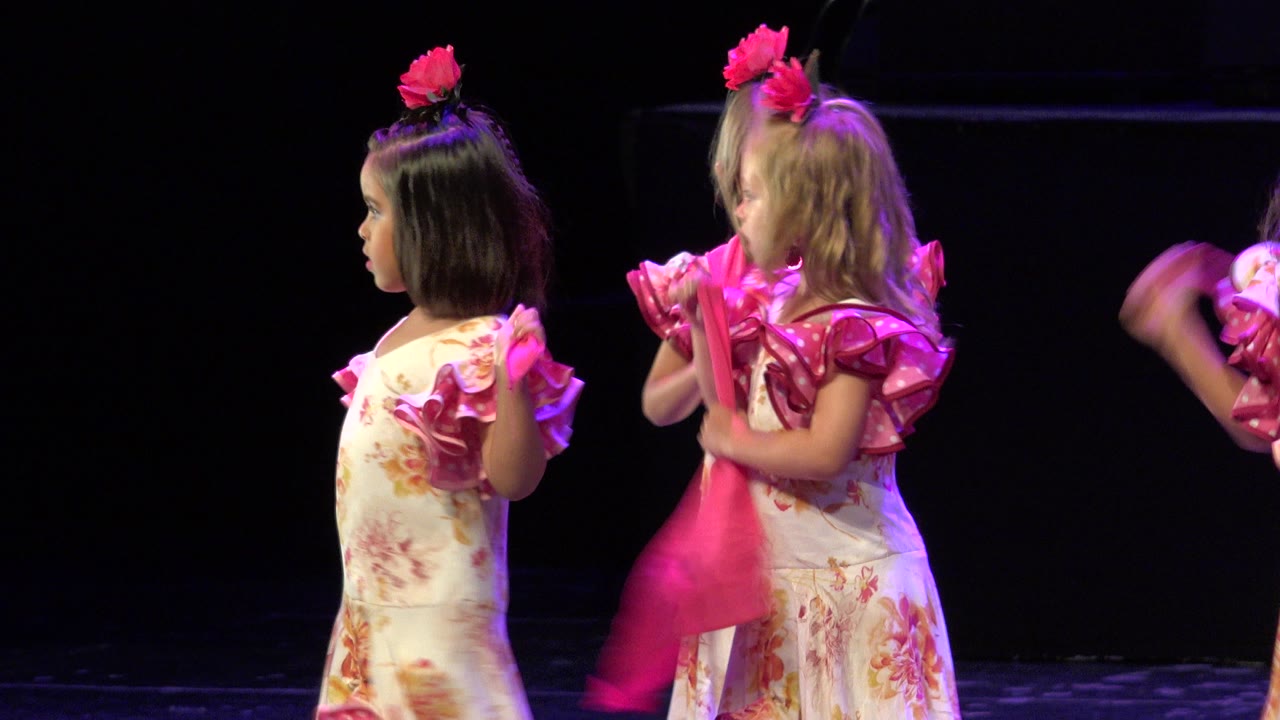 Two young girls in matching floral dance costumes stand side by side on a dark stage in Cabo San Lucas, Mexico, with a blurred arm entering at the right edge.