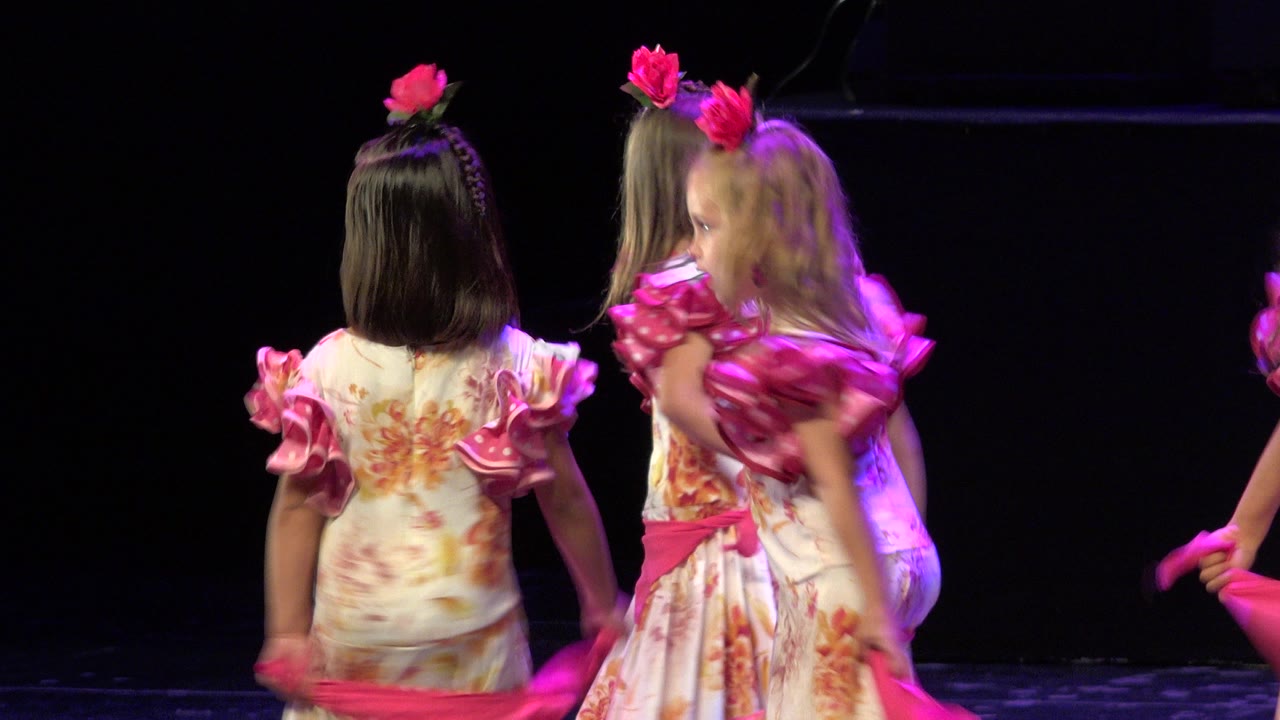 Two little girls in floral dresses stand on a dark stage holding a pink sash.