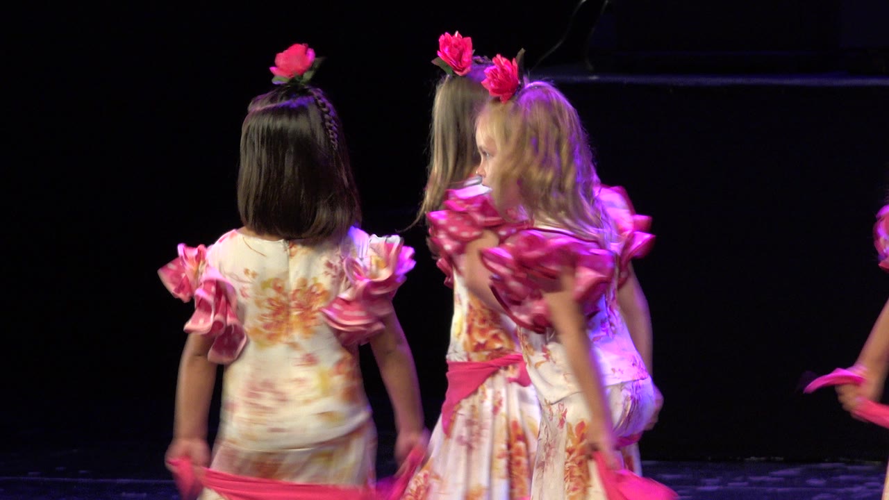 The two girls remain centered with their backs to the camera, their pink hair flowers and sash standing out against the black stage background in Cabo San Lucas.