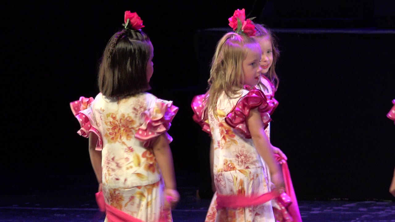 At 0:00 in Cabo San Lucas, Mexico, young girls in floral costumes with pink ruffled sleeves stand close together on a dark indoor stage.